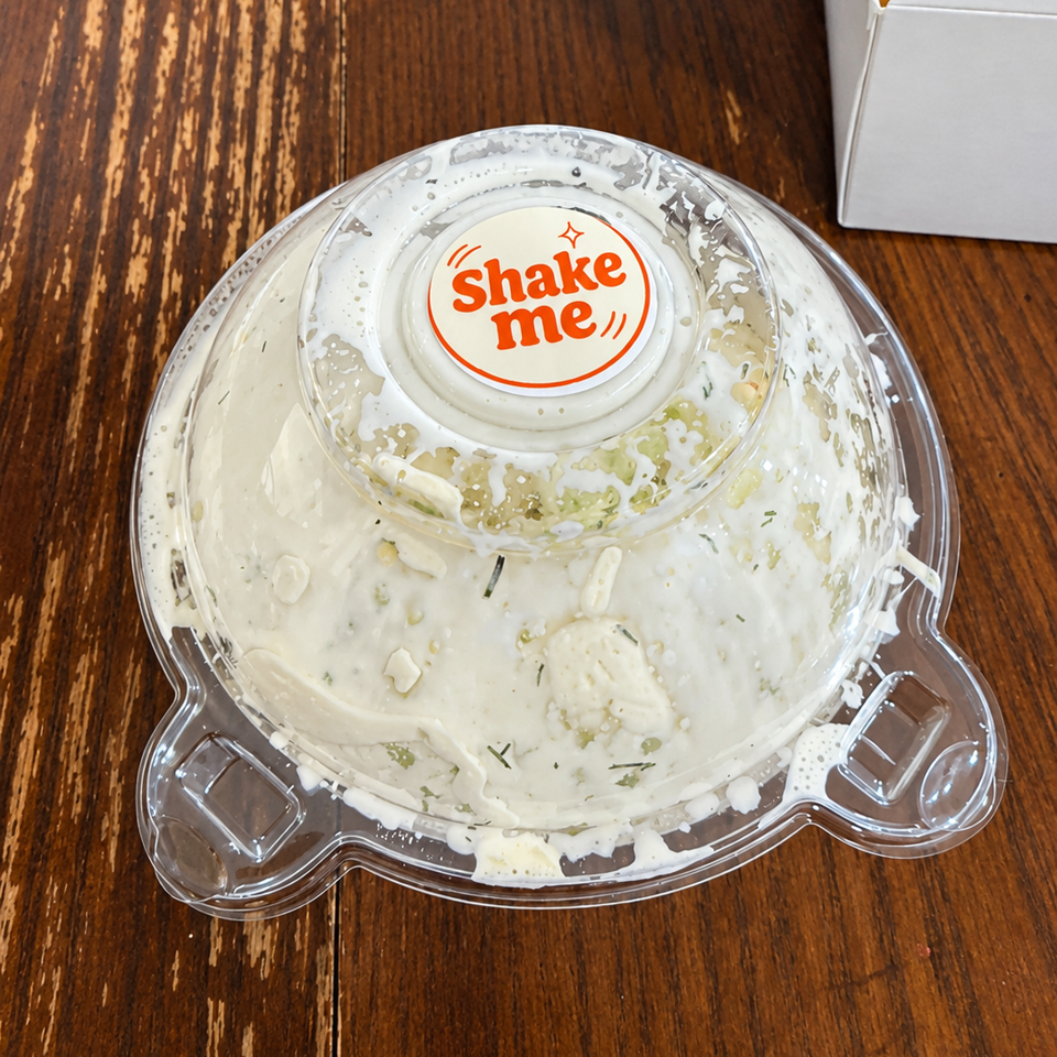 Creamy herb dip in a clear plastic container labeled “Shake me” sits on a rustic wooden table beside a takeout box.